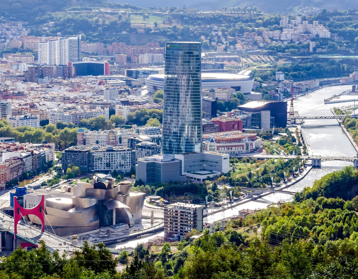  A high-angle view of Bilbao showing the Guggenheim Museum’s sculptural design, a tall modern tower, residential neighborhoods and the Nervión River winding through green hills.