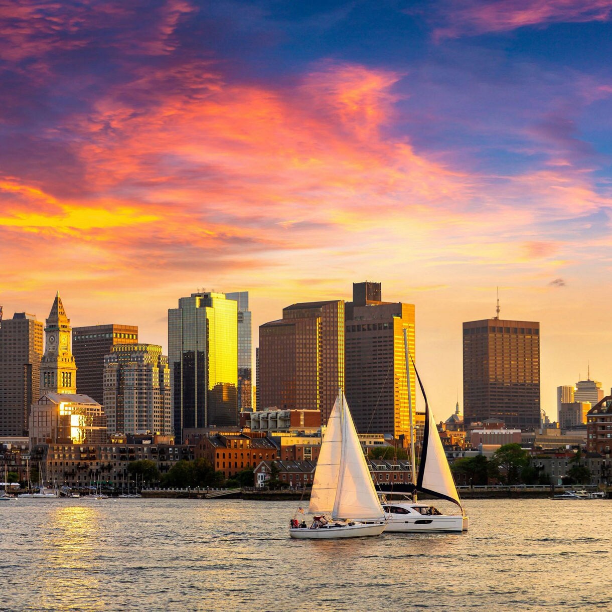A vibrant view of the Boston skyline during a colorful sunset, featuring modern skyscrapers and historic architecture. Sailboats glide across the calm waters in the foreground, adding a serene touch to the urban scene. The sky is painted with hues of orange, pink, and purple, creating a dramatic backdrop.