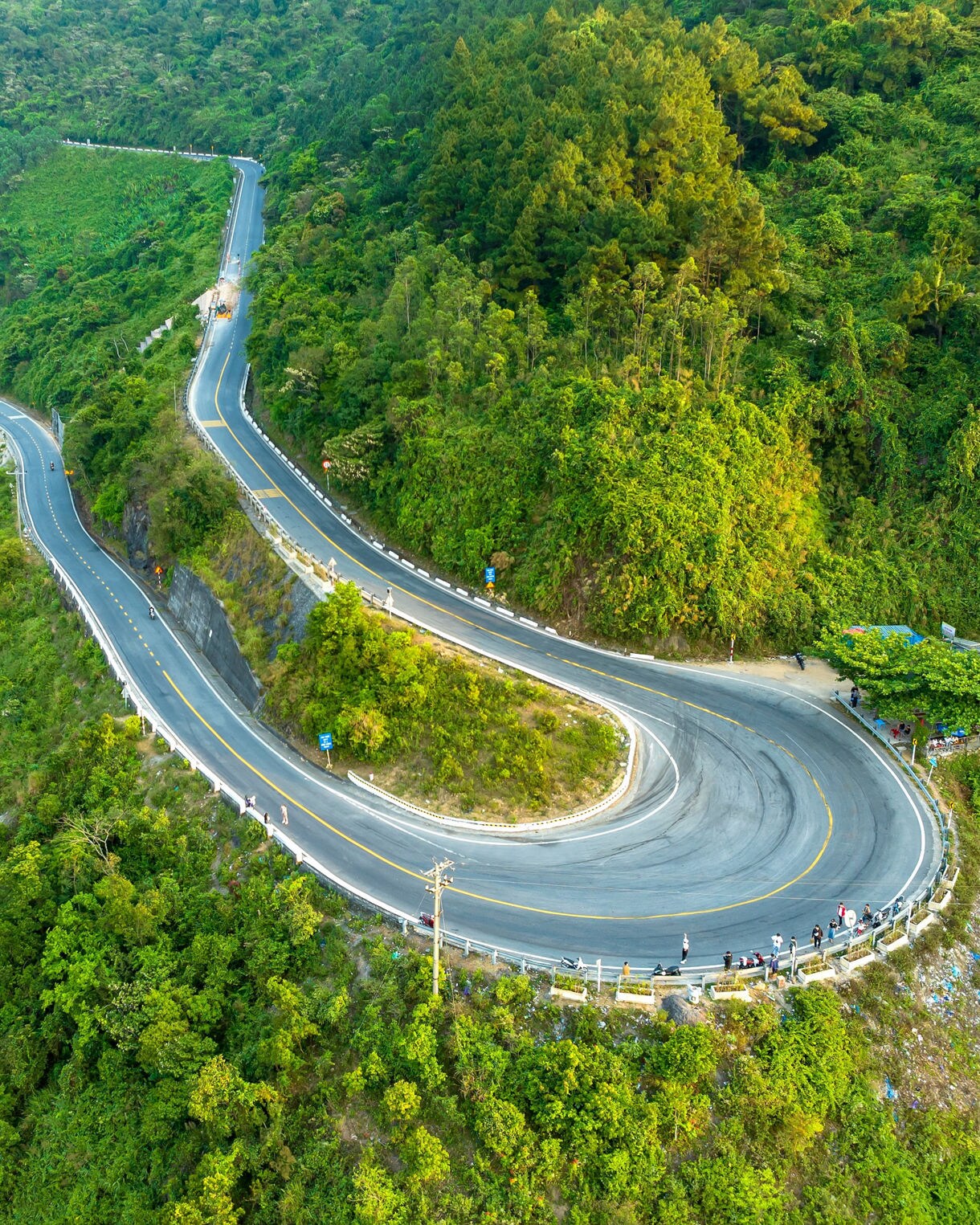 Aerial view of the curving Hai Van Pass road cutting through green mountains in central Vietnam, with vehicles and travelers stopping at scenic viewpoints.