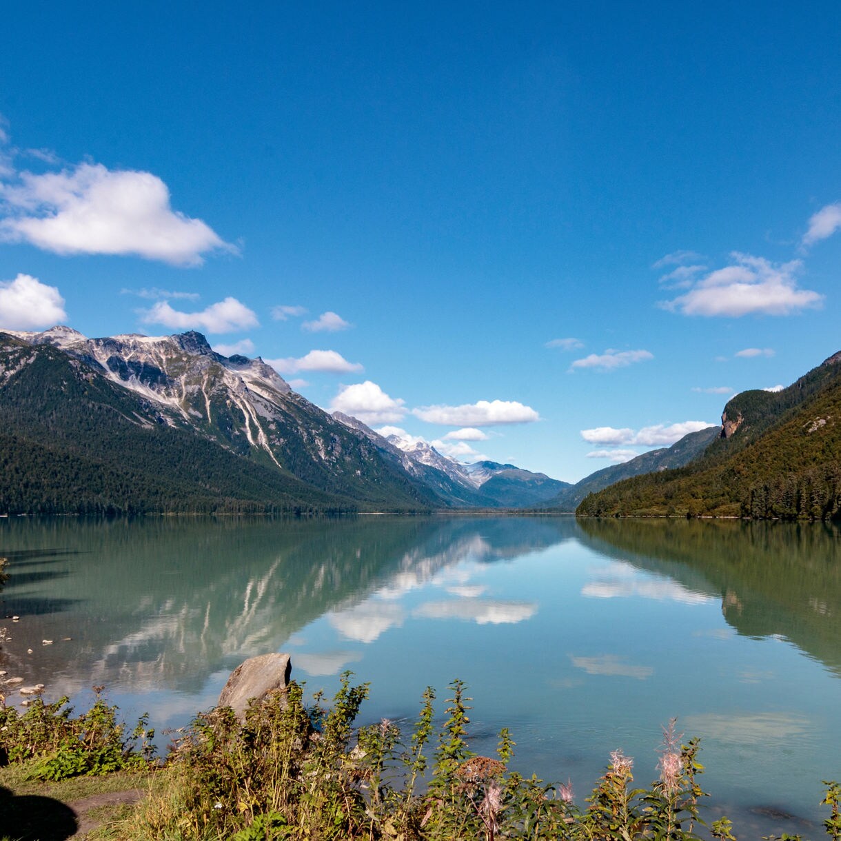 Clear, turquoise waters of Chilkoot Lake reflecting surrounding forested mountains under a bright blue sky with scattered clouds.