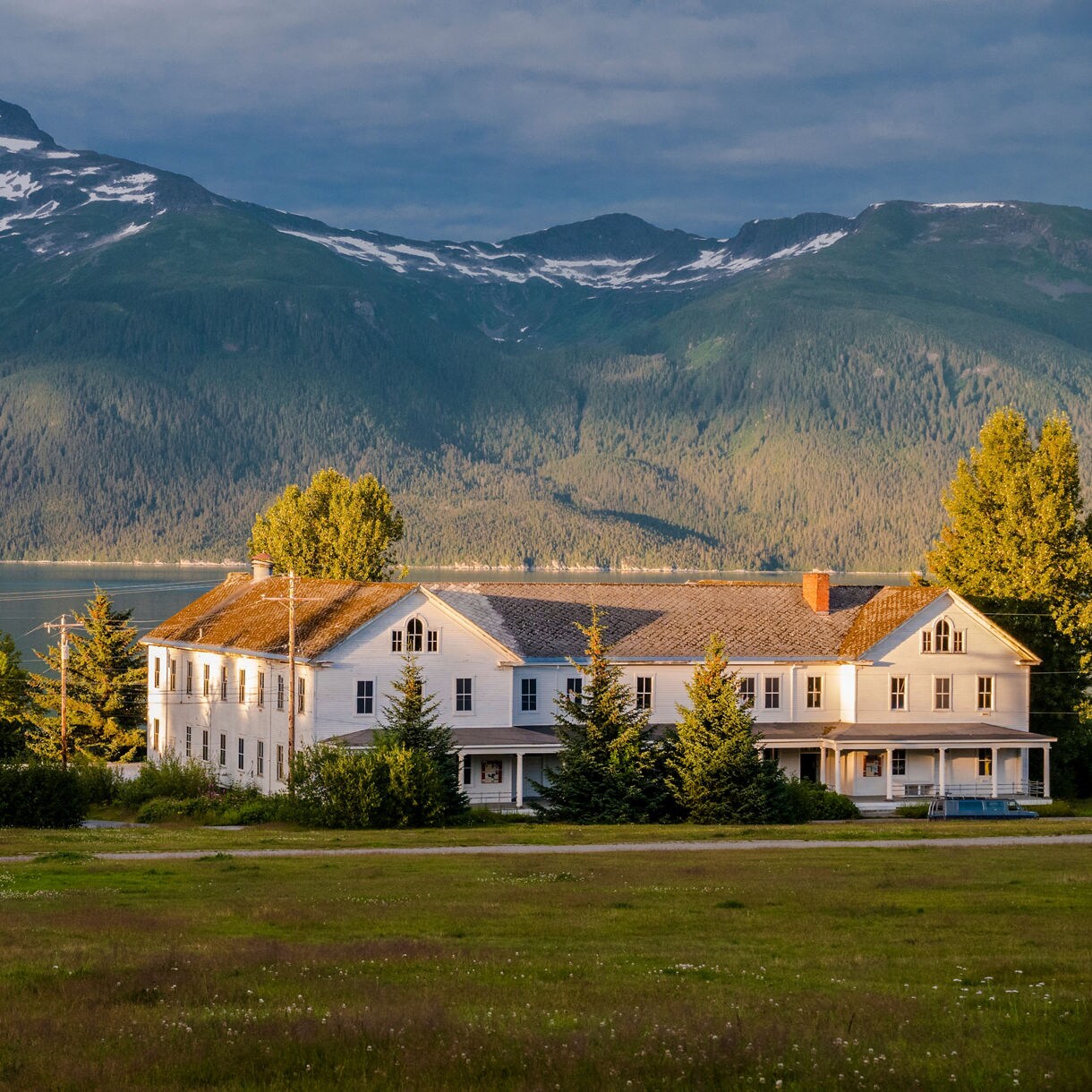 Historic white military building at Fort William H. Seward in Haines, Alaska, with lush green trees and towering mountains in the background.