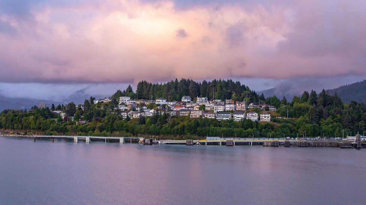 Colorful homes nestled among trees on a hill in Haines, Alaska with a dock in the foreground and dramatic pink and purple clouds overhead at sunset.