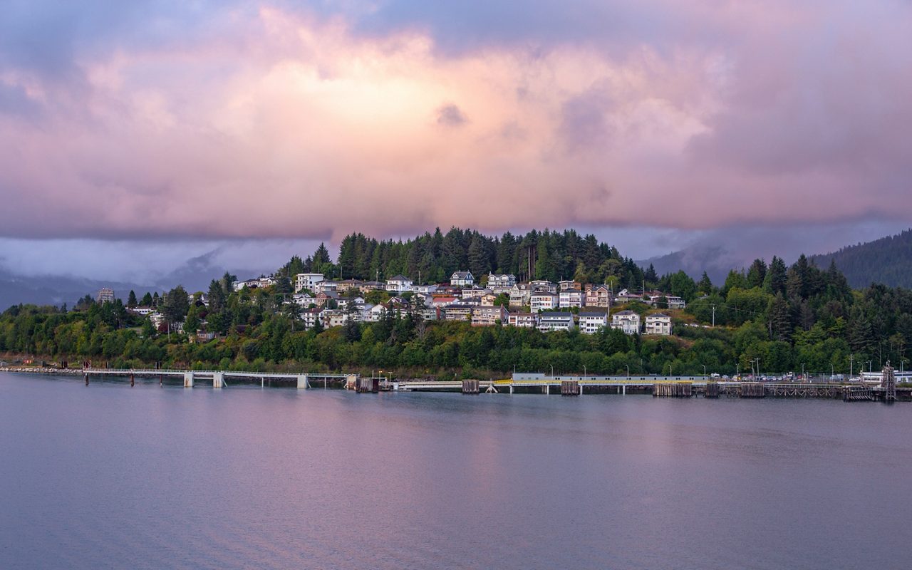 Colorful homes nestled among trees on a hill in Haines, Alaska with a dock in the foreground and dramatic pink and purple clouds overhead at sunset.