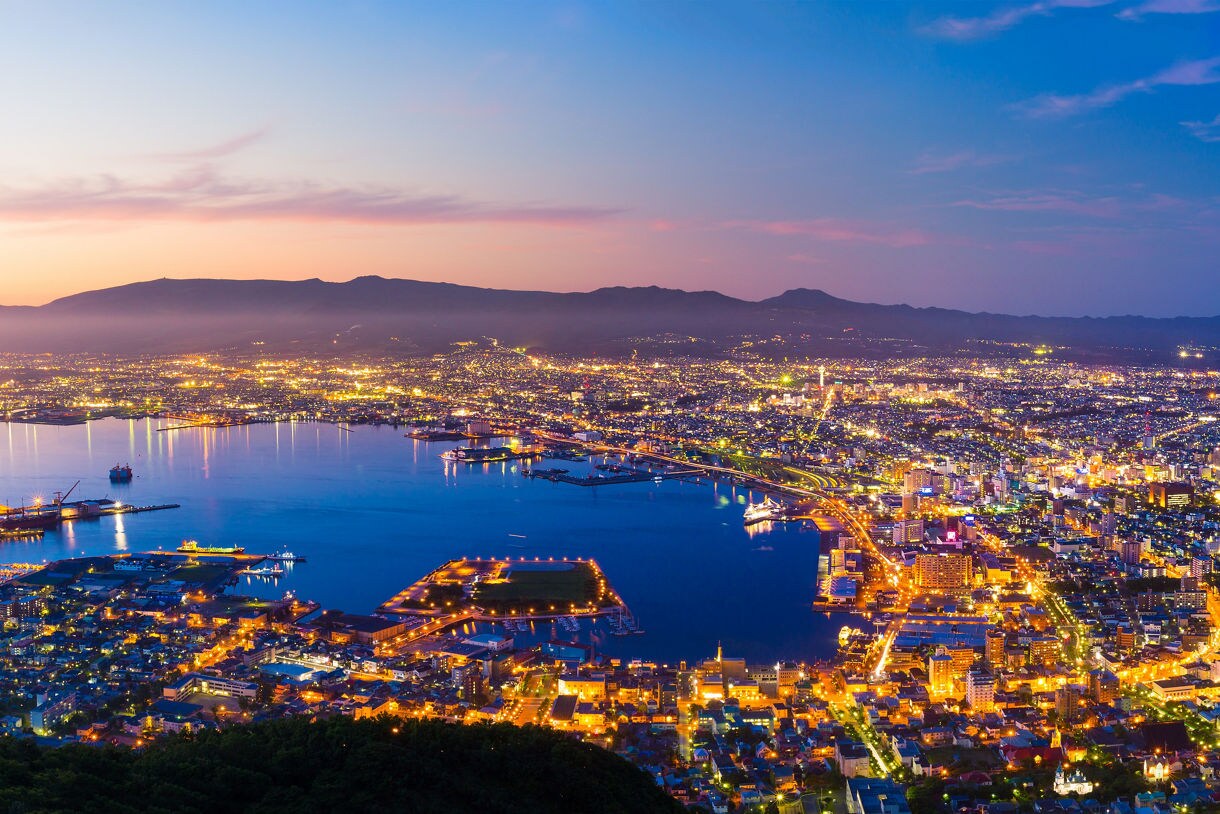 Panoramic twilight view of Hakodate, Japan from above, showing the city illuminated with golden lights along the coastline, a calm bay to the left and a curving shoreline to the right, all framed by distant mountains and a fading blue and pink sky.