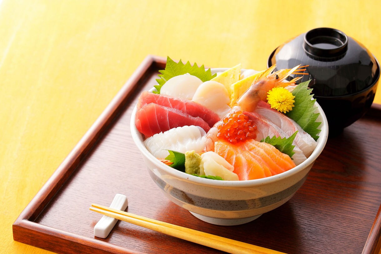 A colorful bowl of kaisendon, a Japanese seafood rice dish, featuring fresh slices of tuna, salmon, squid, scallop, shrimp and fish roe, garnished with shiso leaves, wasabi and a yellow flower, served on a wooden tray with chopsticks and a black lacquered soup bowl.
