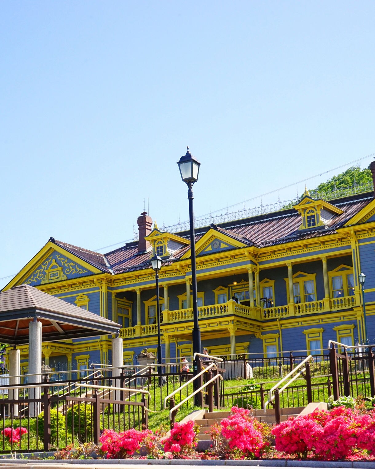 The Former Hakodate Public Hall, a striking Western-style building painted blue with yellow trim, stands beneath a clear blue sky, surrounded by lush trees and blooming pink flowers, with steps and railings leading up through a well-kept garden.