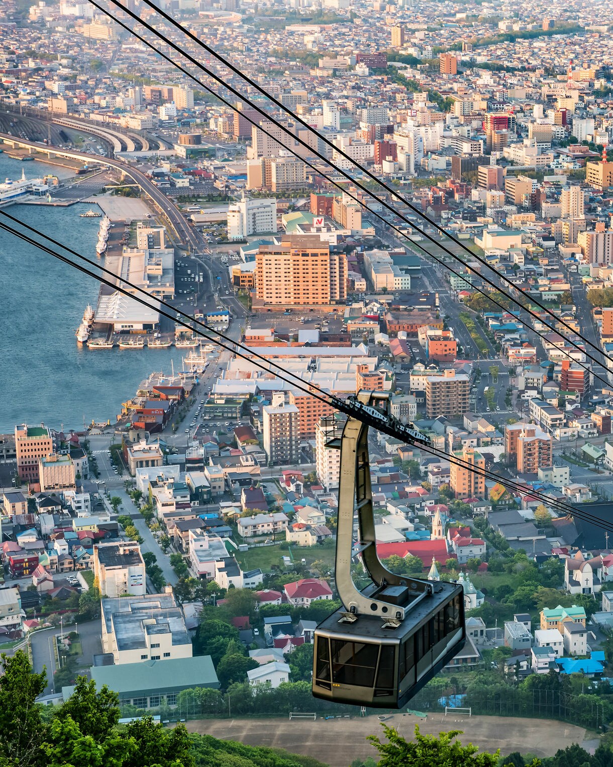 A cable car of the Hakodate Ropeway ascends above the cityscape of Hakodate, Japan, offering a sweeping view of densely packed buildings, a curving coastline and the harbor below, with greenery in the foreground and soft afternoon light casting long shadows.