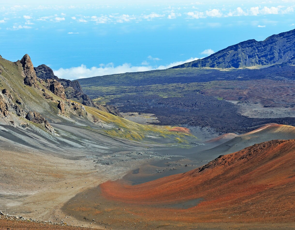 Panoramic view of Haleakalā Crater with red volcanic slopes, misty clouds and distant mountain ridges under a clear blue sky.