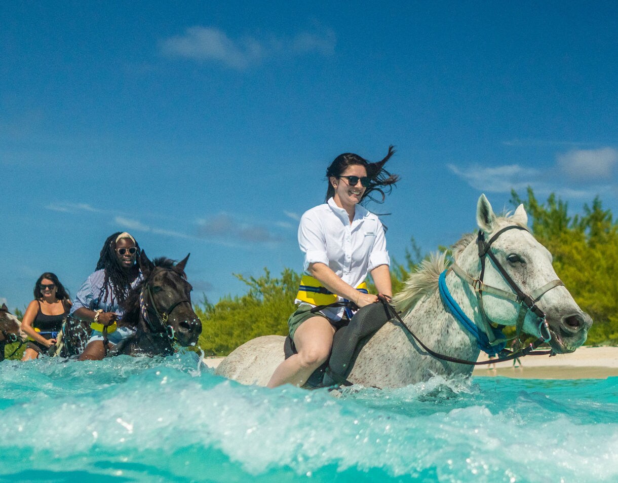 A group of people ride horses through shallow, clear turquoise water near a sandy beach.