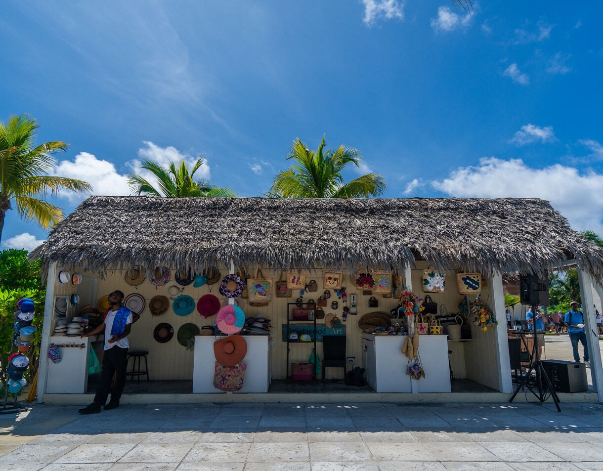 Thatched-roof beach shop with palm trees, colorful hats and decorative items displayed on walls, and visitors browsing inside