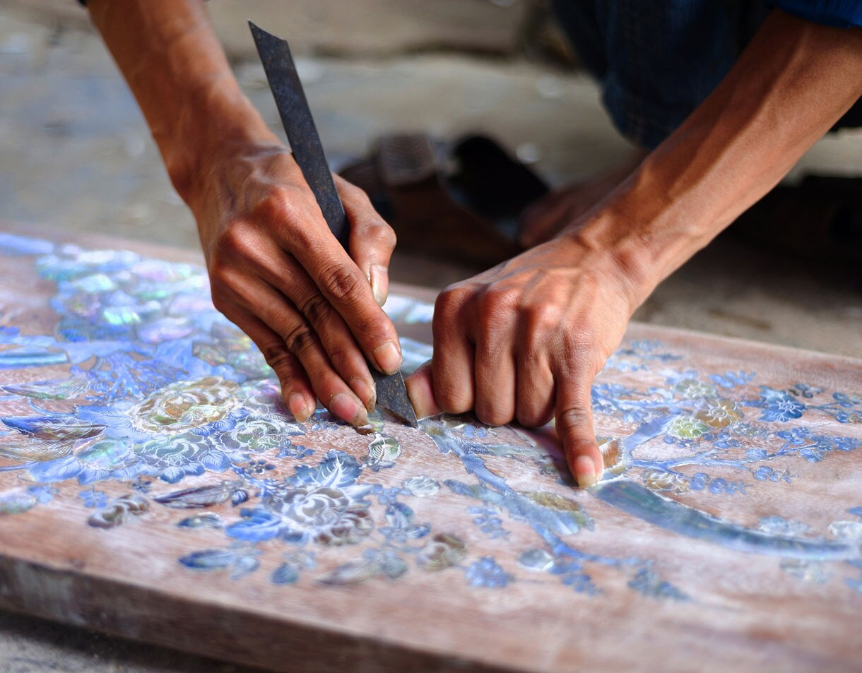 Close-up of a craftsman’s hands carving detailed floral designs with mother-of-pearl inlay on a wooden surface in Vietnam.