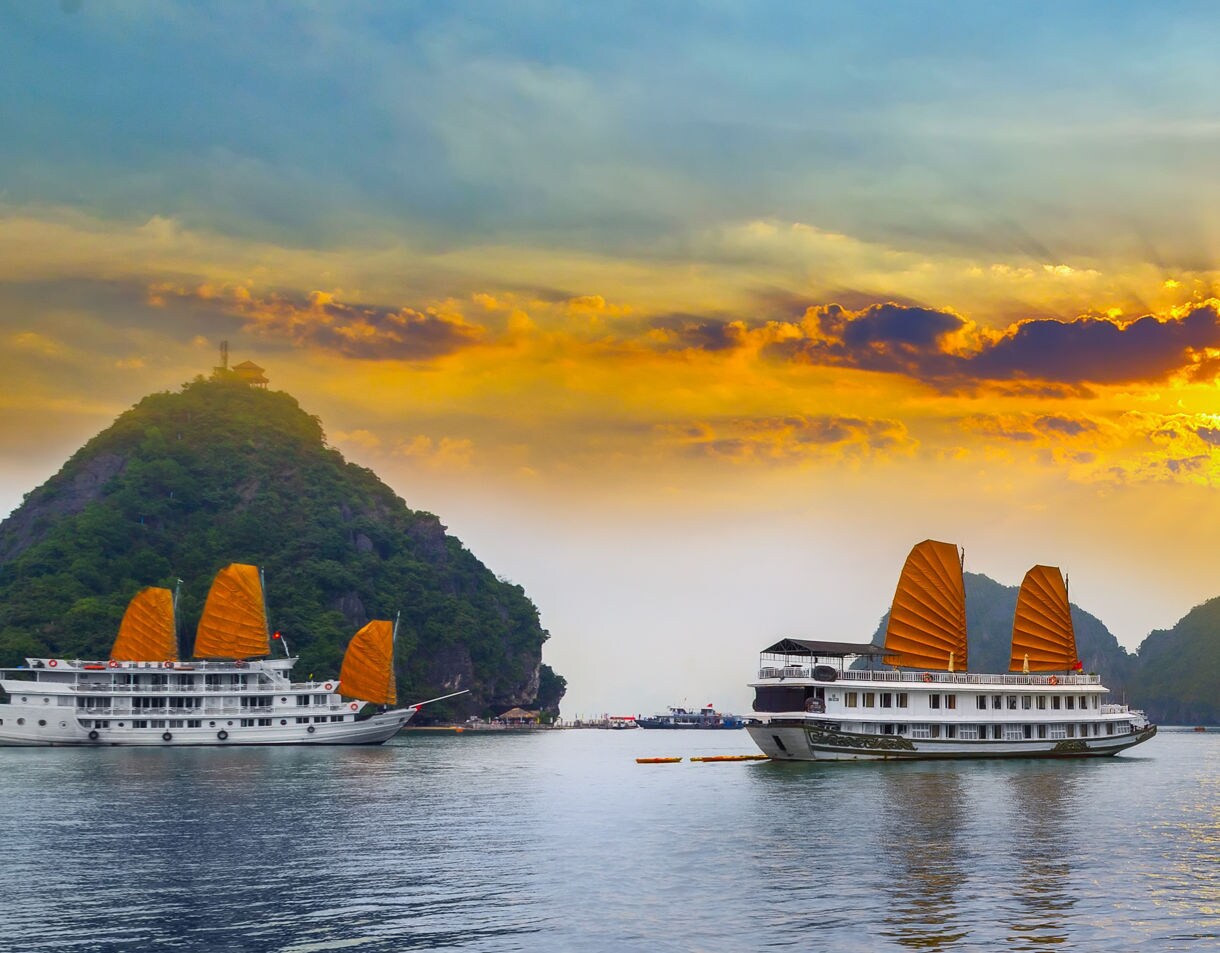 Two white cruise boats with orange sails floating on Hạ Long Bay, Vietnam, beneath a dramatic sunset sky with limestone hills in the background.