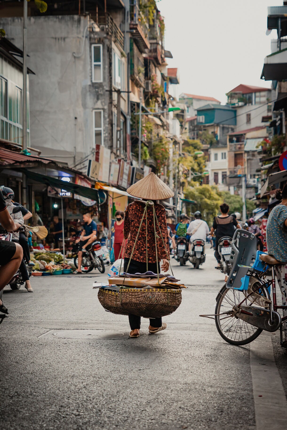 A Vietnamese street vendor in a conical hat carries goods in hanging baskets along a busy street in Hanoi’s Old Quarter, surrounded by motorbikes, shops and market stalls.