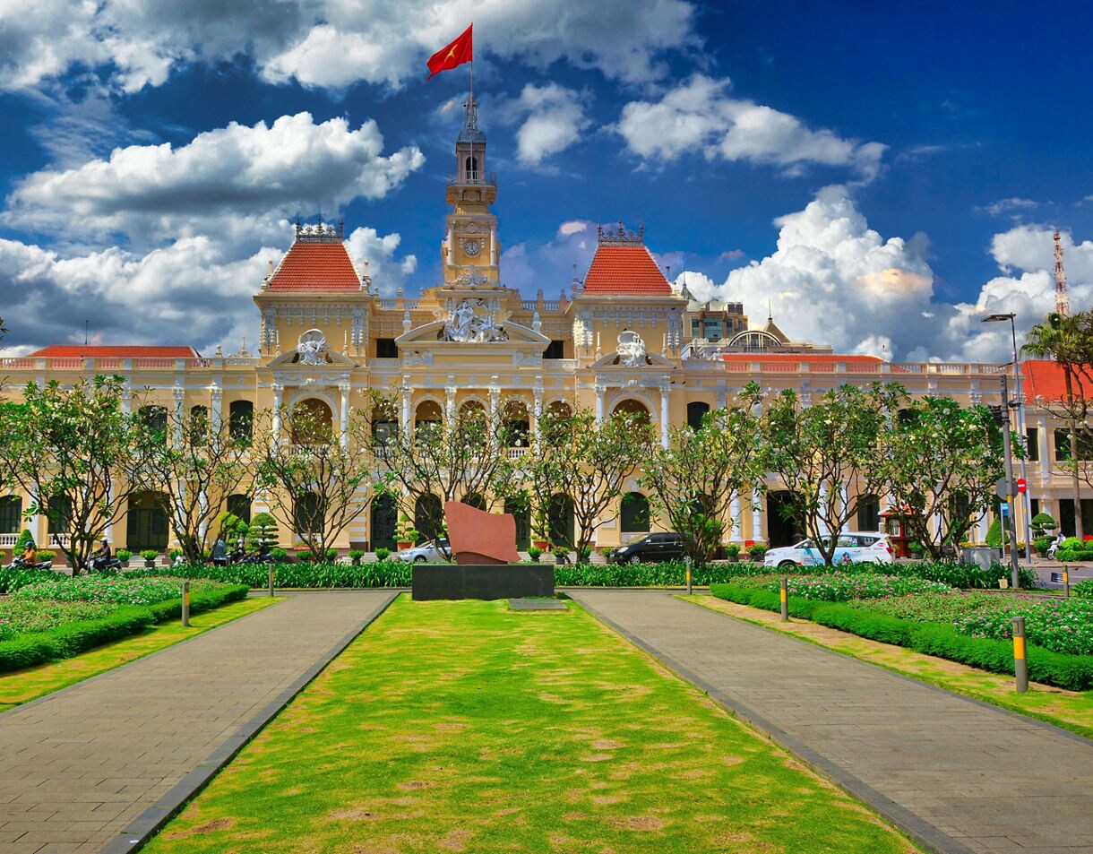Ho Chi Minh City Hall in Vietnam, a grand French colonial building with yellow walls, red-tiled roofs, ornate carvings and a tall clock tower flying the Vietnamese flag.