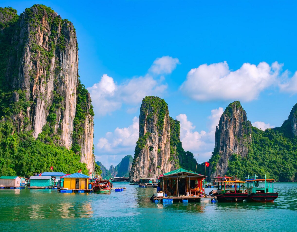 Colorful floating houses and boats on the emerald waters of Hạ Long Bay, Vietnam, surrounded by steep limestone cliffs covered in greenery.