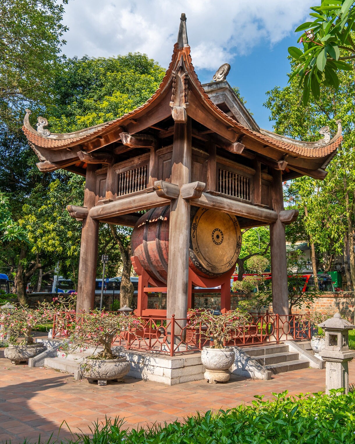 Wooden pavilion with a large ceremonial drum at the Temple of Literature in Hanoi, Vietnam, surrounded by lush gardens, stone lanterns and bonsai pots.