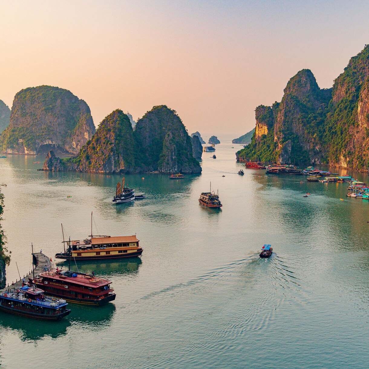 Boats and junks scattered across Hạ Long Bay in Vietnam, surrounded by steep limestone karsts covered in greenery during a calm, hazy sunset.