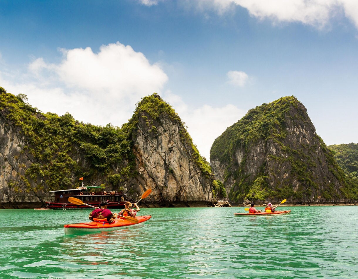 Tourists kayaking in bright orange kayaks across the green waters of Hạ Long Bay, Vietnam, with steep limestone karsts covered in vegetation in the background.