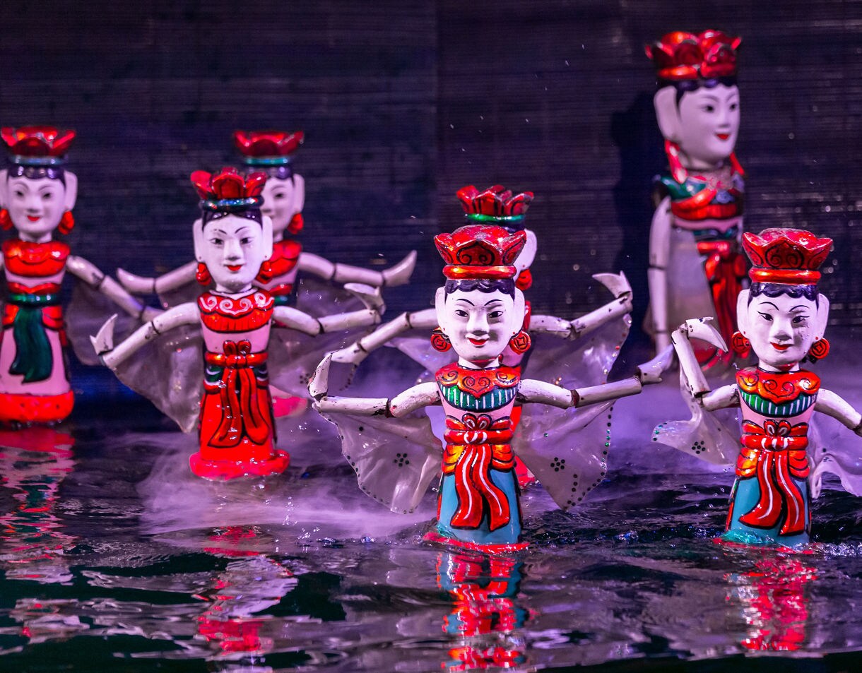 Wooden water puppets dressed in red and green costumes performing on a water stage in Vietnam, with mist rising around them.