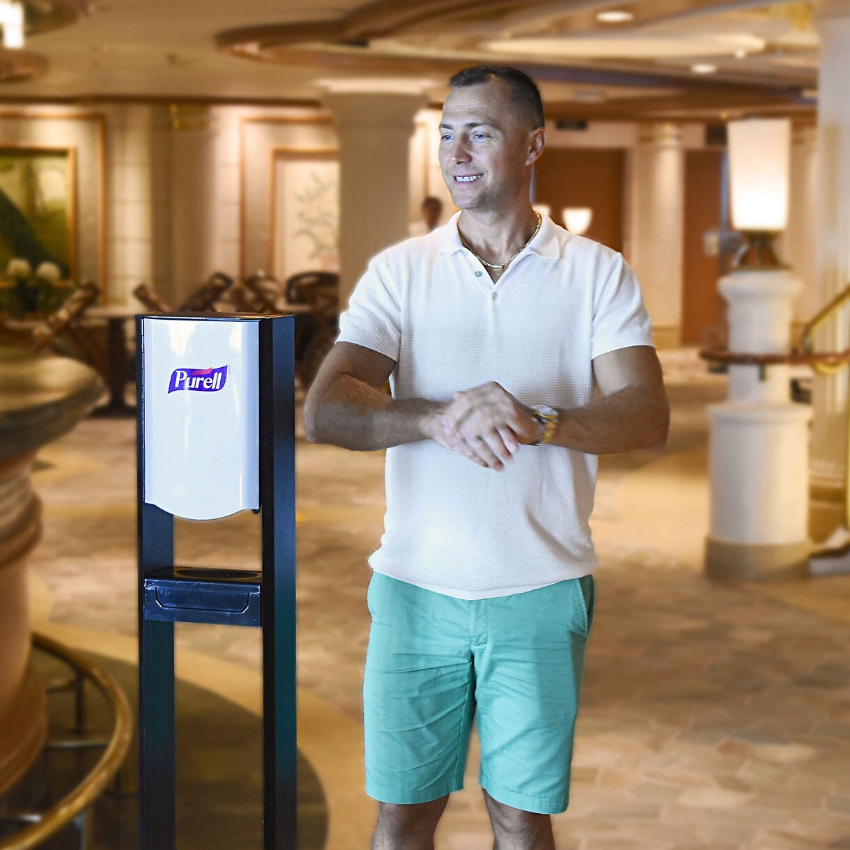 A man stands in front of a Purell hand sanitizer dispenser, applying sanitizer to his hands. The scene takes place in a well-lit hotel lobby or upscale public area, featuring elegant decor and ambient lighting. The man is casually dressed in a white polo shirt and green shorts, and the Purell logo is clearly visible on the dispenser. The atmosphere is clean and welcoming, emphasizing hygiene and safety.