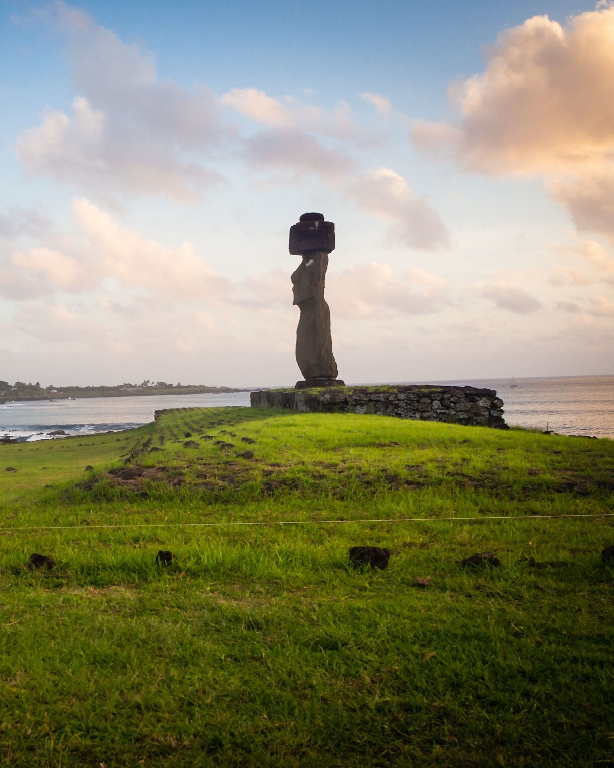 Single moai statue with a red pukao standing on a stone platform near the ocean at Hanga Roa, surrounded by green grass under a pastel sunset sky.