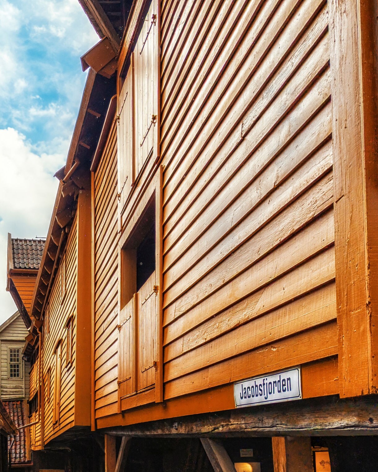 Narrow alleyway in Bergen’s Bryggen district with historic wooden Hanseatic buildings, one painted orange with a “Jacobsfjorden” sign.