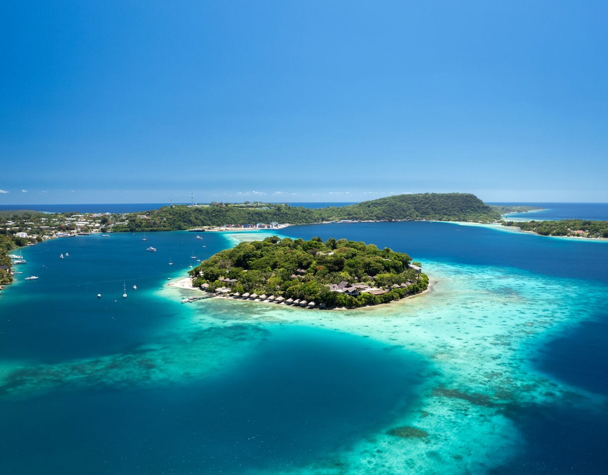 Aerial view of Iririki Island surrounded by bright turquoise water and deep blue lagoon, with nearby coastline and forested hills in the background.