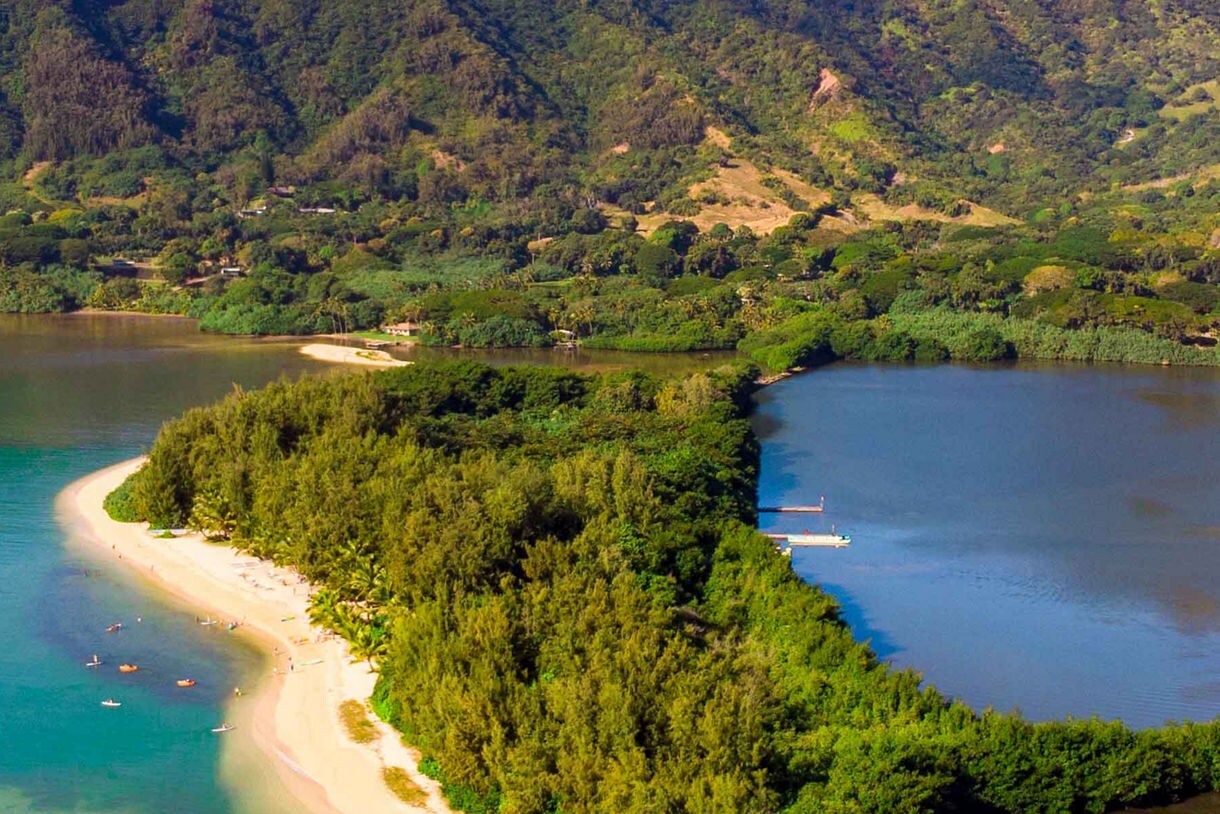 Aerial view of a curved white sand beach with turquoise and deeper blue waters, bordered by dense tropical vegetation and rolling green hills.