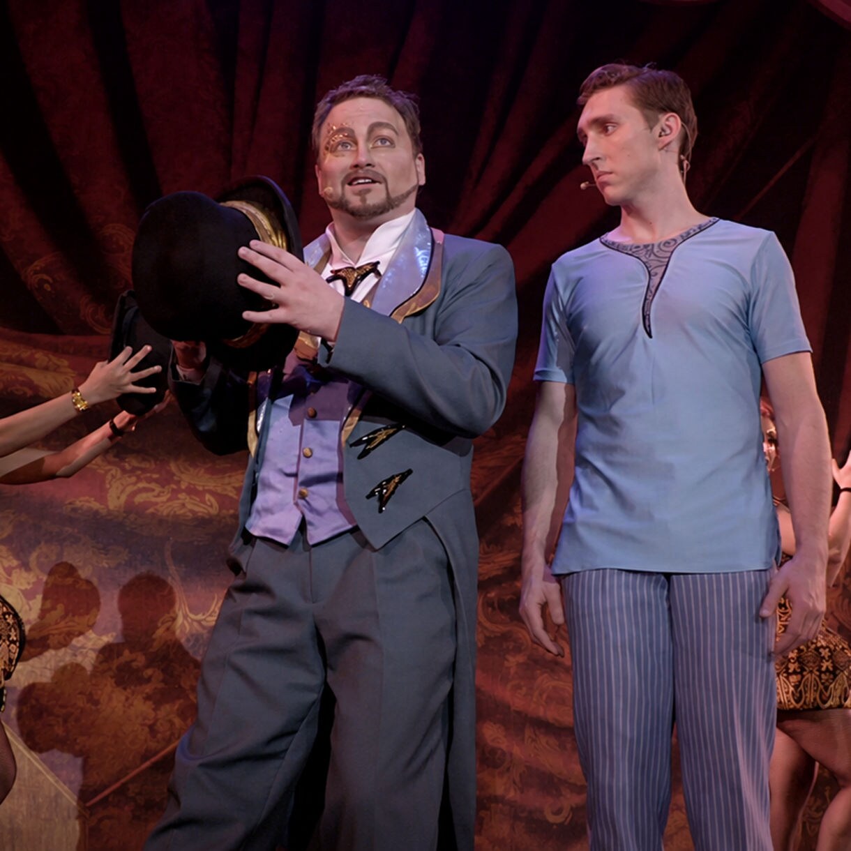 Three performers stand on a theater stage in front of a large, ornate curtain. The central actor in a suit holds a black top hat while addressing the audience. A female dancer in a patterned costume reaches toward the hat, and a male performer in casual stage attire stands to the right. Warm stage lighting and rich fabric textures create a classic, dramatic atmosphere.