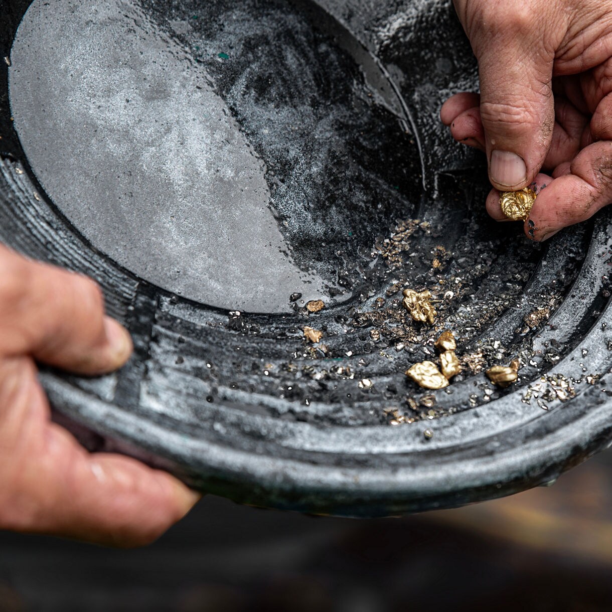 Close-up of hands panning for gold with a metal tray full of gravel and water over train tracks and rocky ground.