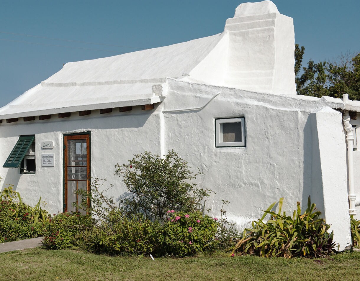 Small whitewashed stone chapel with a sloped roof, green shutters and flowering plants around the entrance under a clear sky.