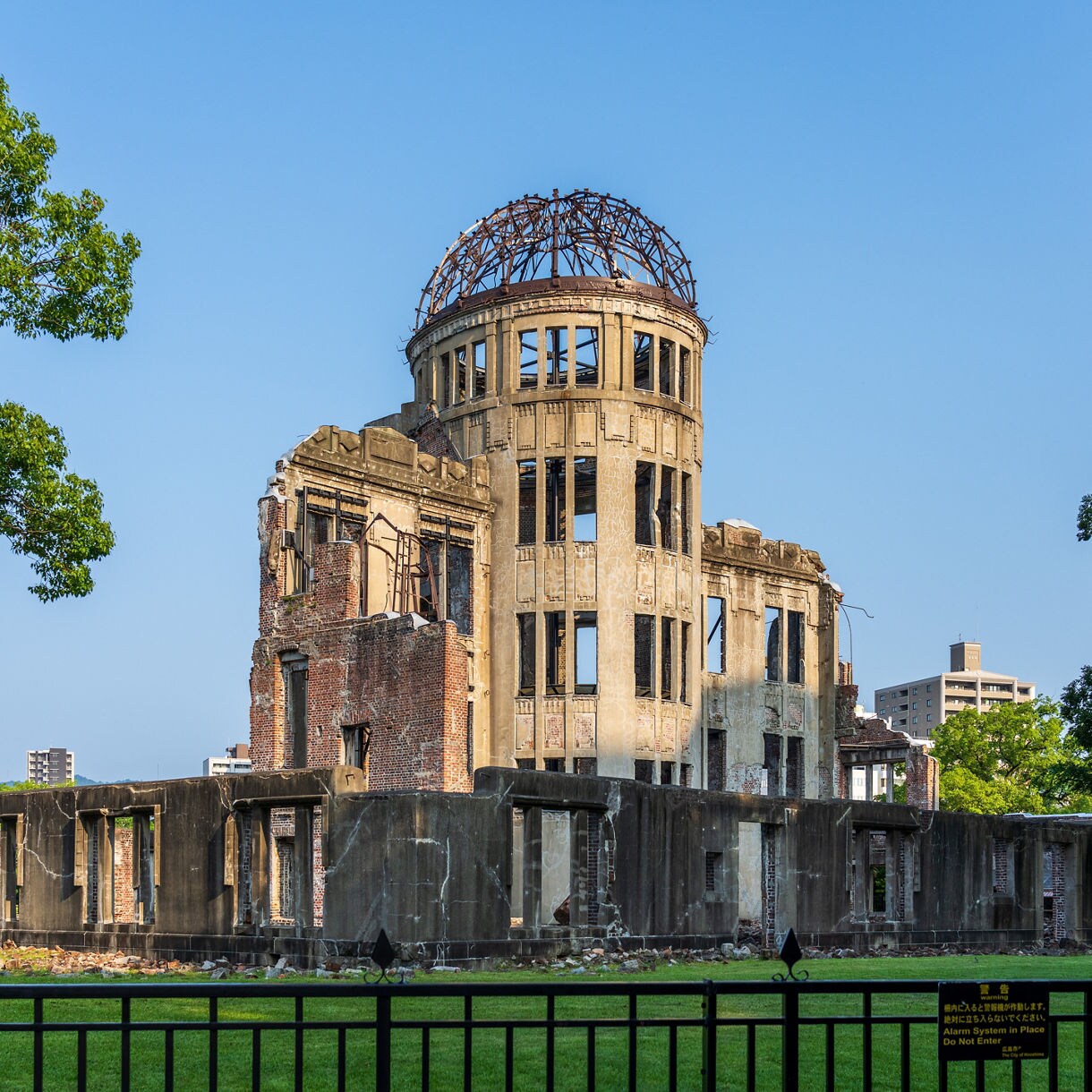 The Genbaku Dome in Hiroshima, Japan, a preserved ruin from the atomic bombing, with its skeletal dome and brick walls surrounded by green grass, trees and city buildings in the background.