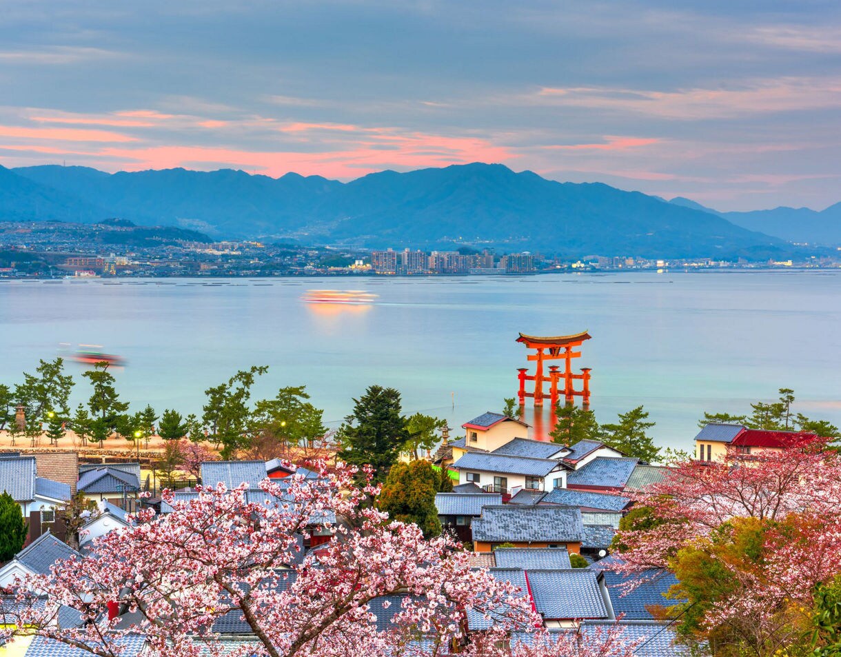 Iconic red Torii Gate of Itsukushima Shrine standing in shallow water near Miyajima Island, with colorful sunset clouds and distant mountains in the background.