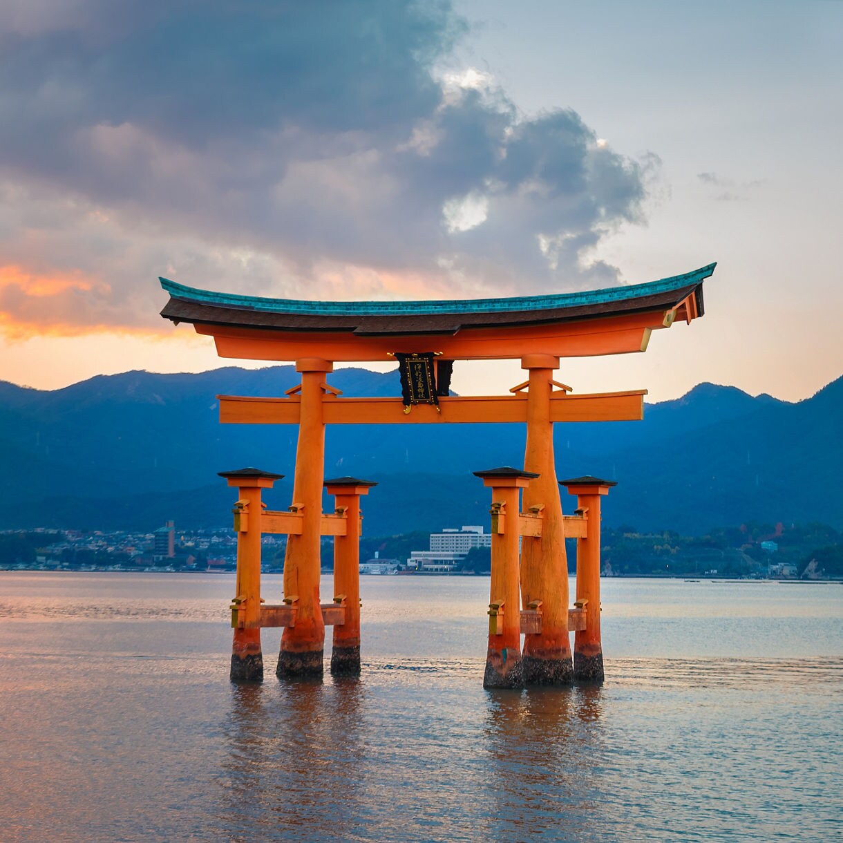 The large red-orange Torii Gate of Itsukushima Shrine standing in shallow water with sunset-lit clouds and dark mountain silhouettes in the background near Hiroshima, Japan.