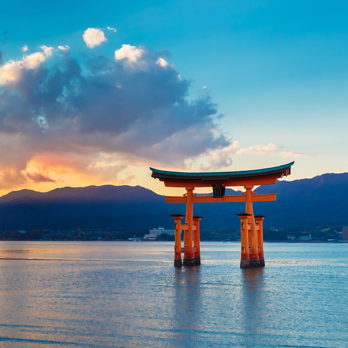 Vermilion torii gate standing in shallow water with calm reflections, backed by distant mountains and a colorful sunset sky over the bay.