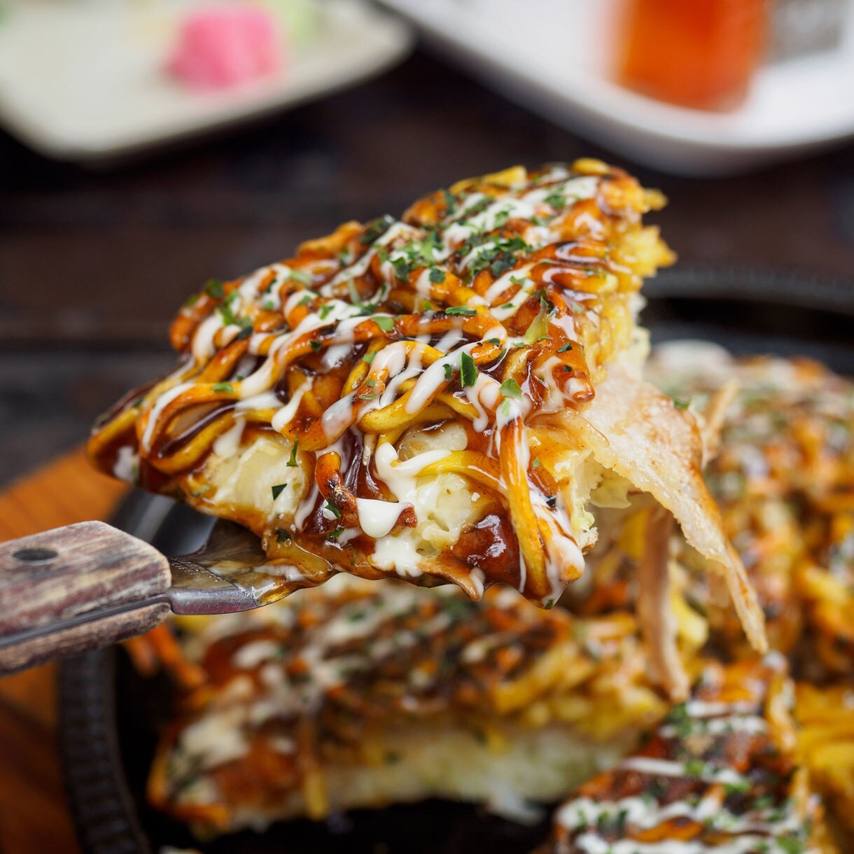 Close-up of a slice of Hiroshima-style okonomiyaki being lifted from a hot plate, topped with drizzled sauces, mayonnaise, seaweed and savory ingredients.