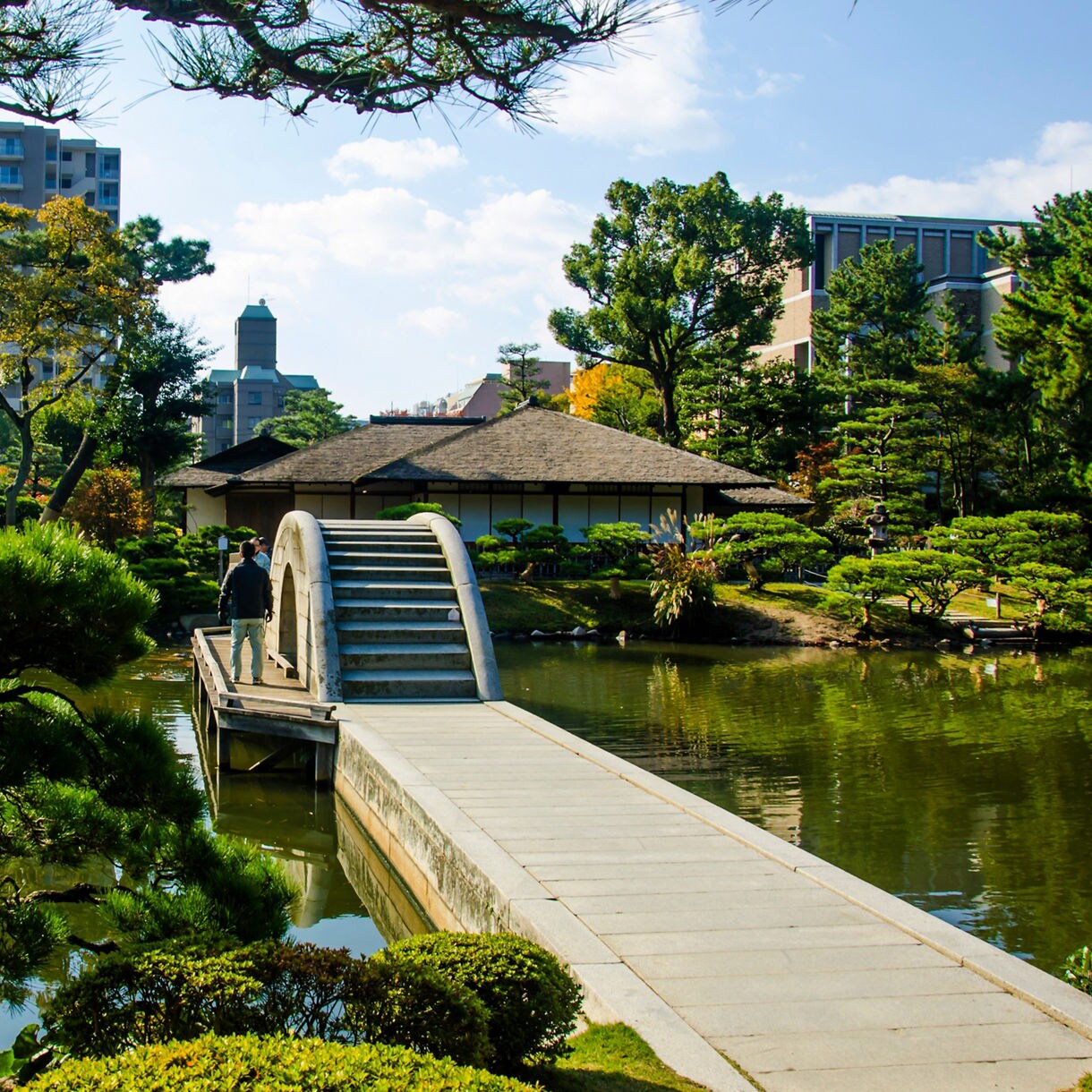Traditional Japanese arched stone bridge over a pond in Hiroshima’s Shukkeien Garden, surrounded by manicured trees, autumn foliage and historic-style buildings