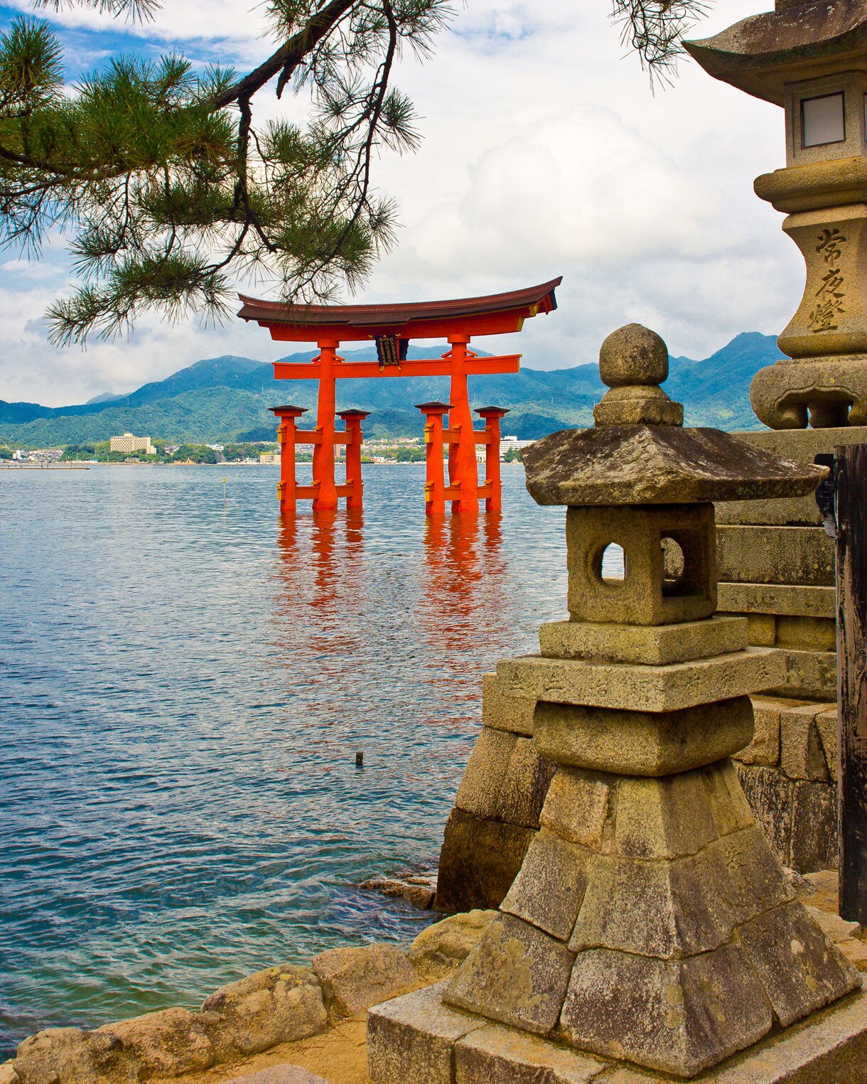 Scenic view of the red Torii Gate of Itsukushima Shrine standing in the water, framed by stone lanterns and pine tree branches.