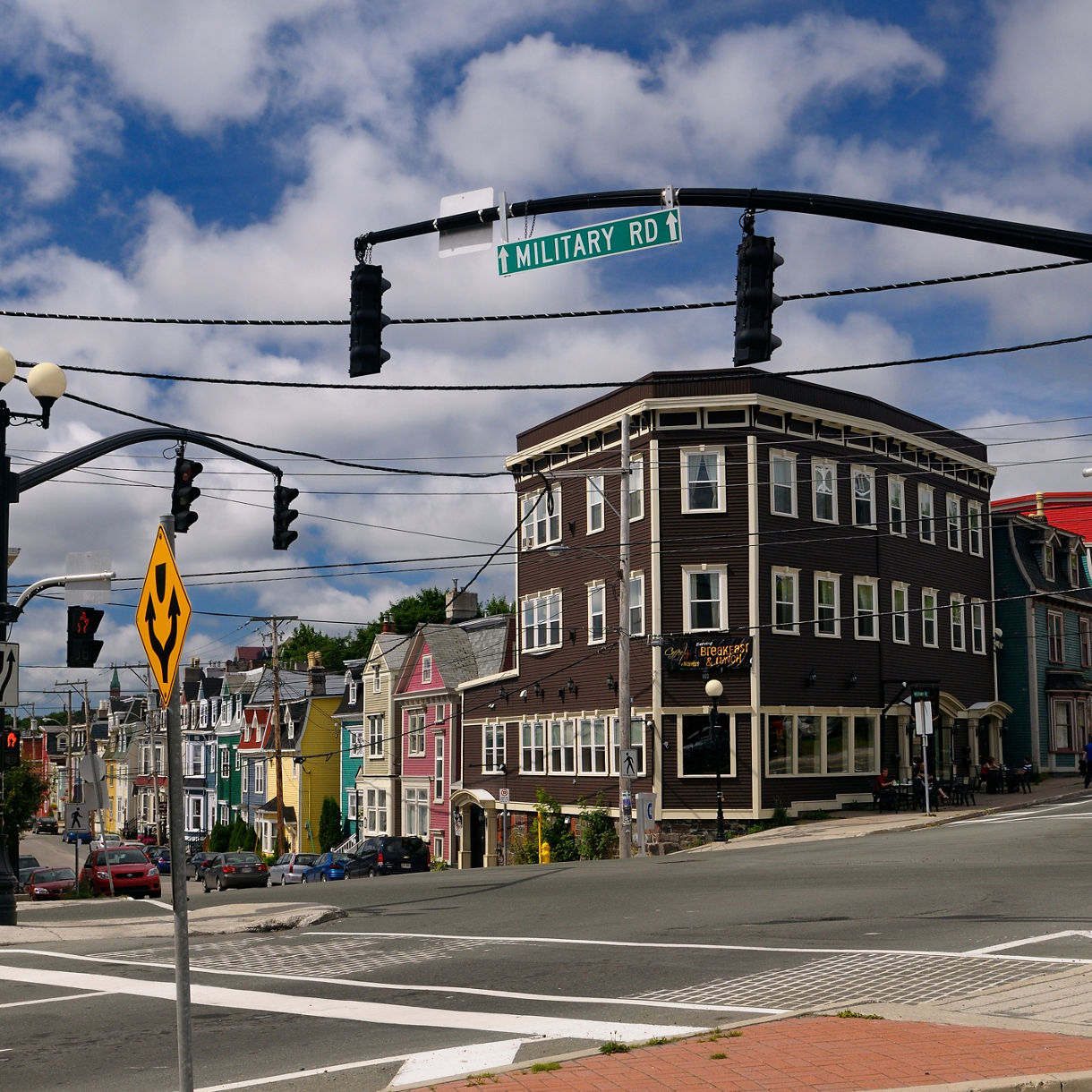 A street corner in St. John’s, Newfoundland, featuring tall multicolored wooden row houses, a dark brown corner building, traffic lights, street signs and overhead power lines beneath a partly cloudy sky.