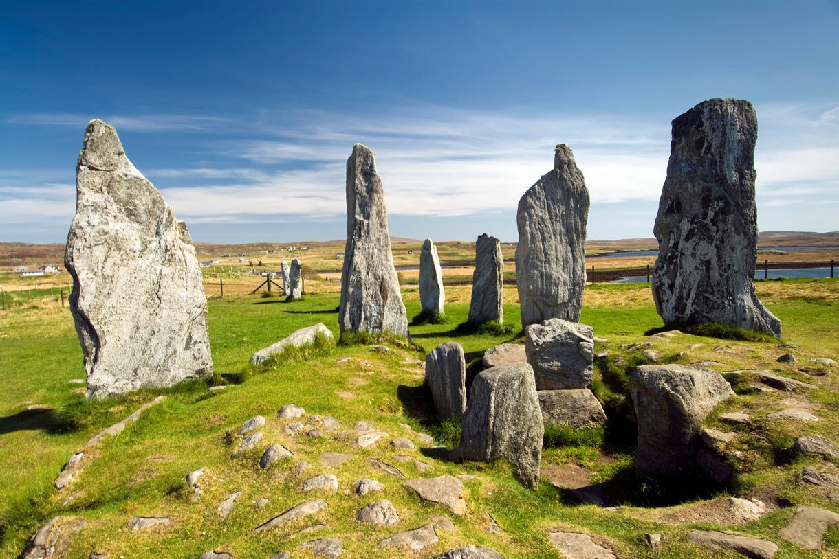 Standing stones arranged in a circular formation on a grassy plain in the Isle of Lewis, with tall weathered monoliths casting shadows under a bright blue sky and rolling moorland stretching into the distance.