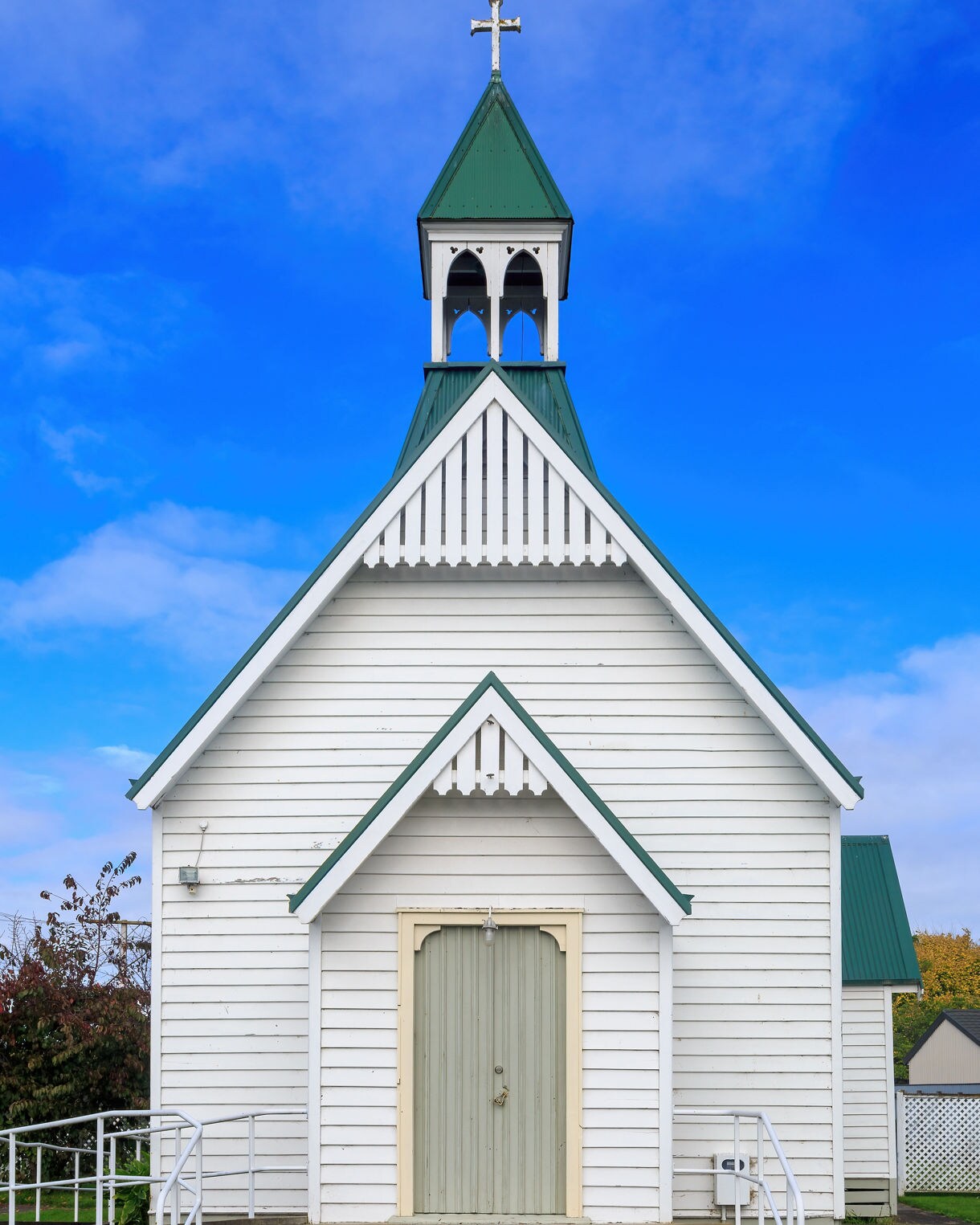 A small white wooden church with a green roof and steeple, viewed from the front, set beneath a vivid blue sky.