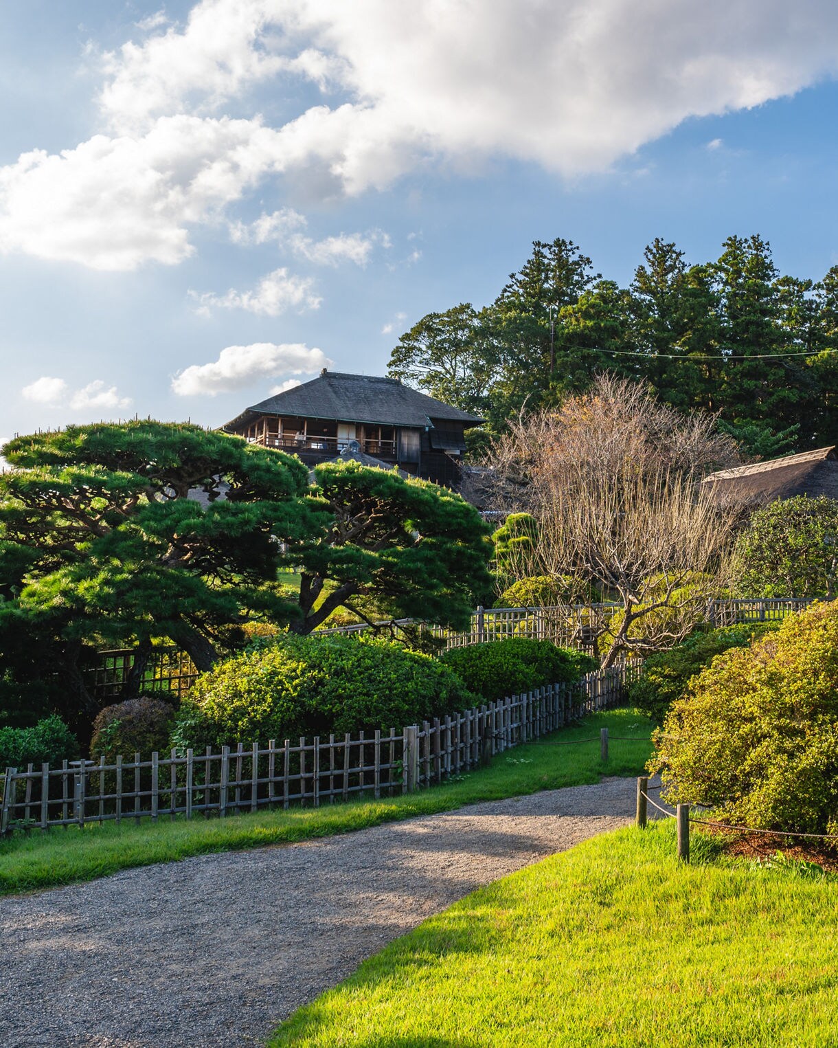 Traditional wooden buildings nestled among manicured trees and lush greenery at Kairakuen Garden under a partly cloudy sky.