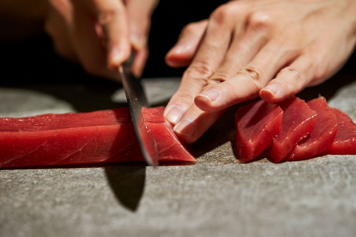 Close-up of hands slicing raw tuna into sashimi pieces with a sharp knife on a cutting board.
