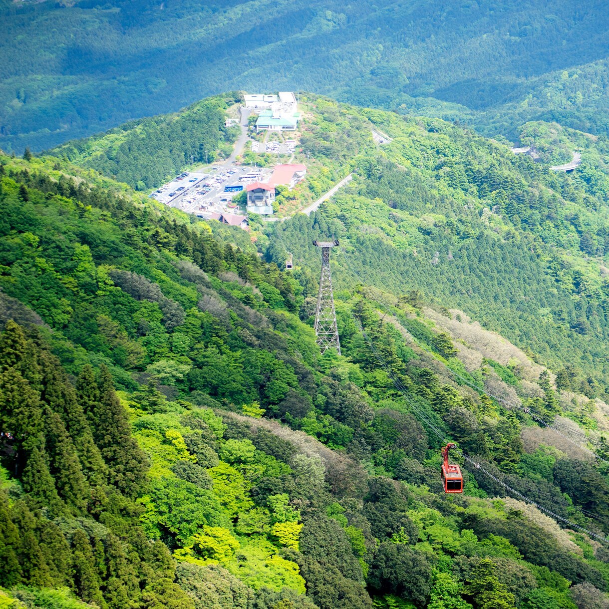 Red cable car traveling over forested mountains near a station on Mt. Tsukuba, surrounded by dense green foliage.
