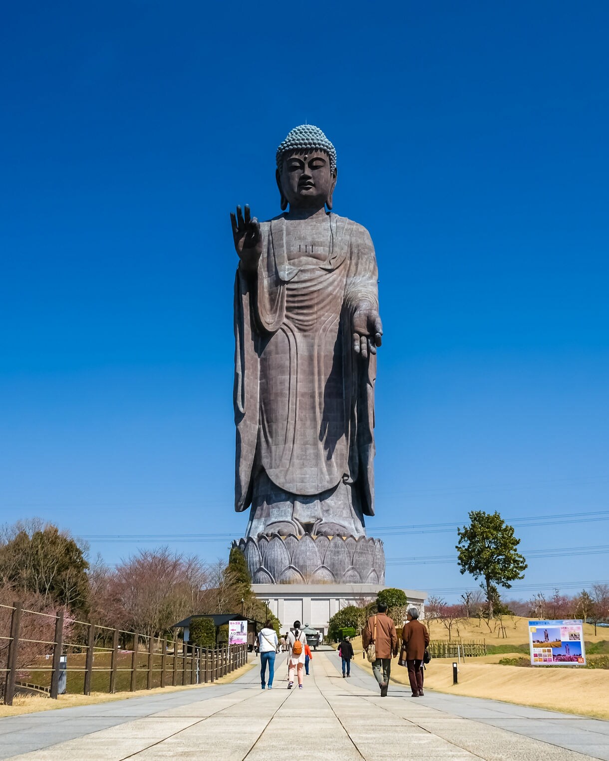 Visitors walk toward the massive bronze statue of Ushiku Daibutsu under a clear blue sky, surrounded by landscaped gardens.