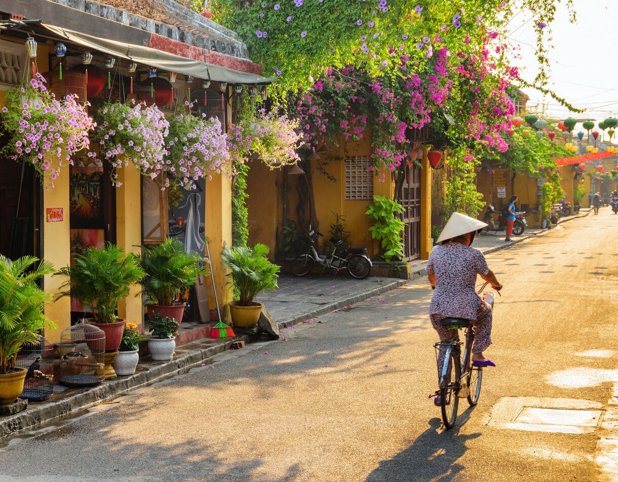 Woman wearing a conical hat rides a bicycle down a quiet Hoi An street lined with yellow buildings, potted plants and hanging flowers in warm morning light.