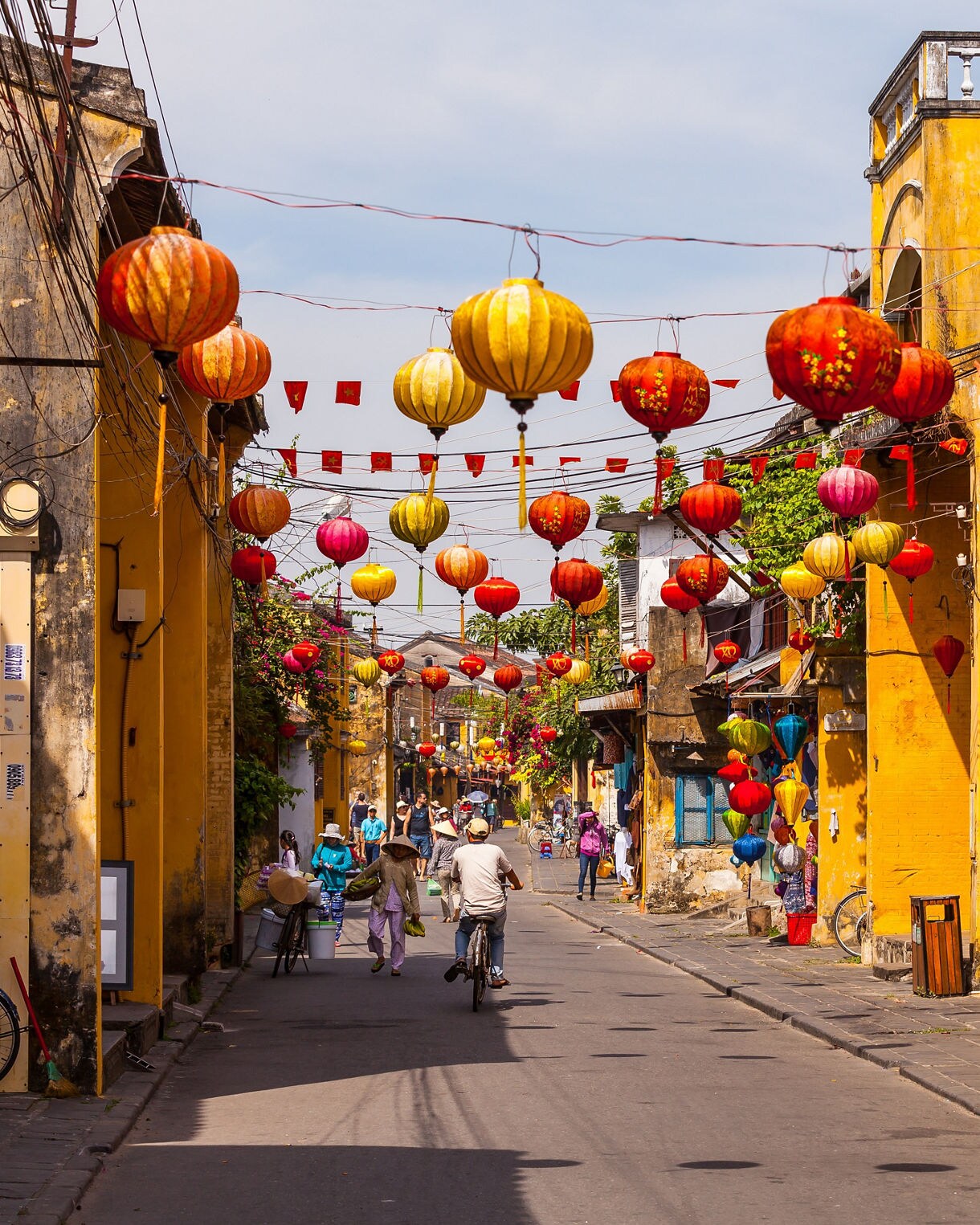 Vibrant street in Hoi An Ancient Town, Vietnam, decorated with colorful hanging lanterns above yellow colonial buildings and cyclists passing by.