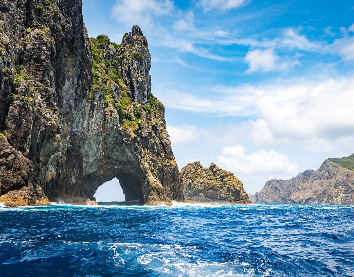 Towering coastal rock formation with a natural arch rising from deep blue ocean waters under a bright, partly cloudy sky.