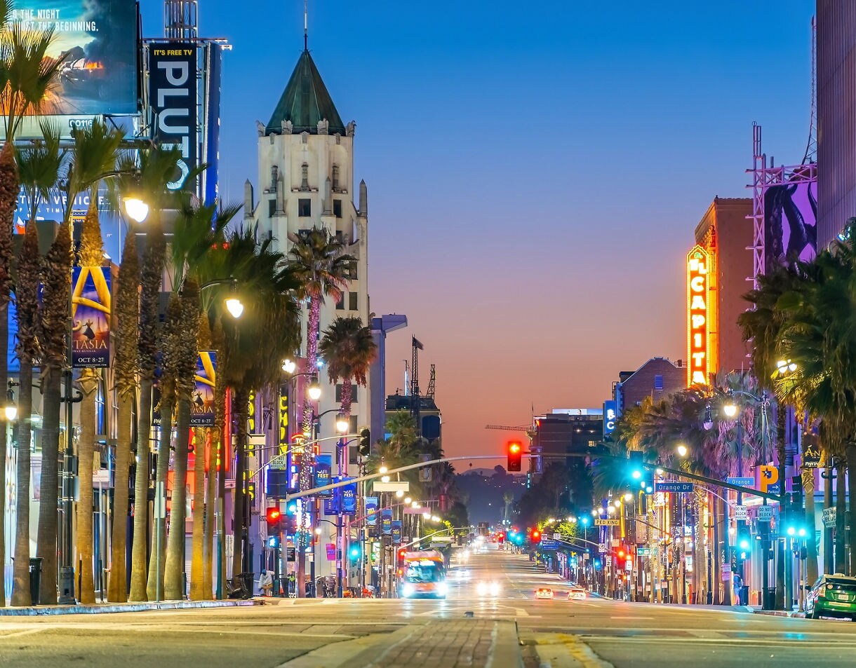 Evening view of Hollywood Boulevard with glowing neon signs, palm trees lining the street and traffic moving beneath a colorful sunset sky.