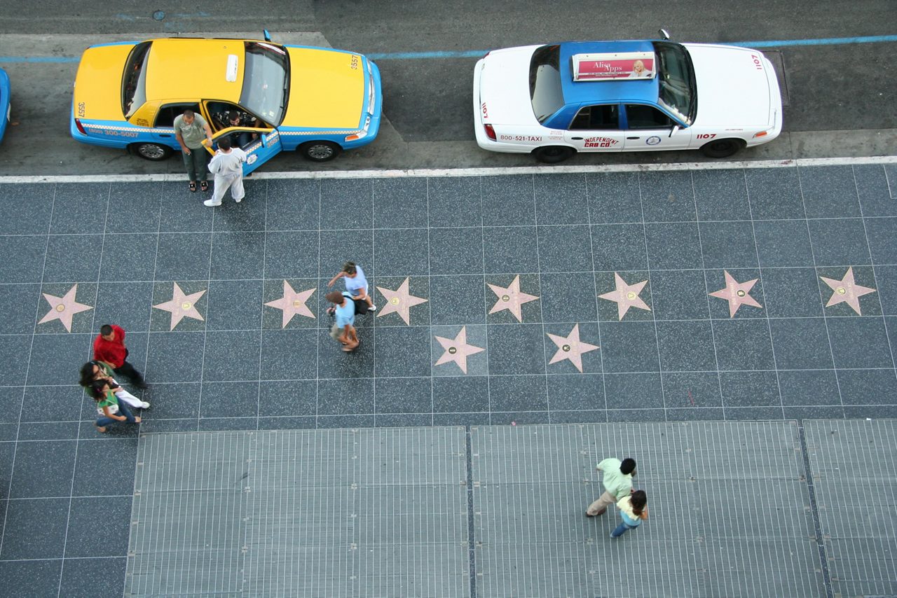 A top-down view of the Walk of Fame on Hollywood Boulevard, showing pedestrians walking past stars embedded in the sidewalk, with taxis parked nearby on the street.